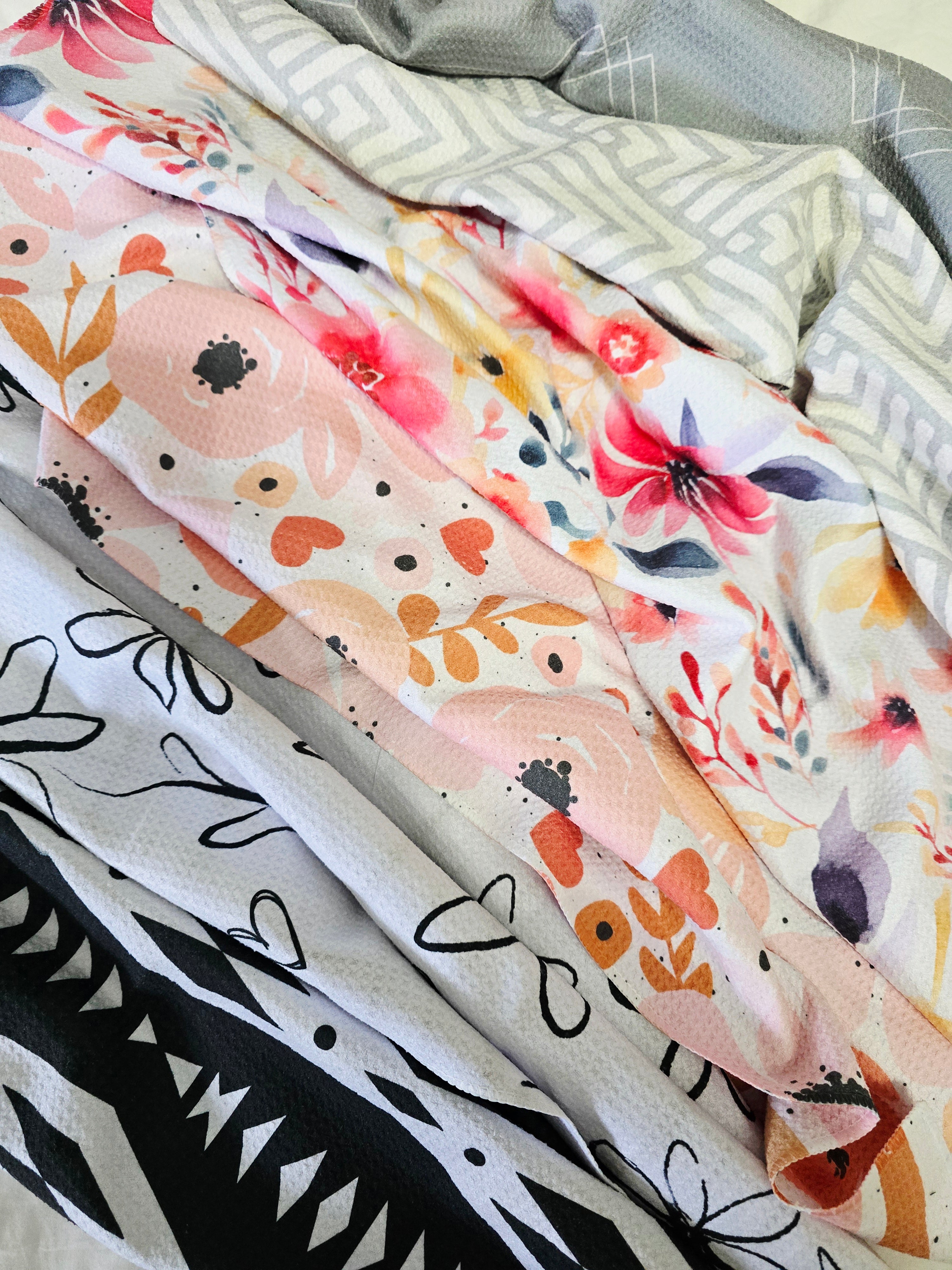 Front view of six colorful microfiber towels laid out right next to eachother to show just a glimpse of the fronts of each. Patterns include black and white geometric triangles, black and white hand drawn daisies, peach, pink and coral hearts and florals, colorful watercolor florals, gray and white geometric triangles and diamonds. 