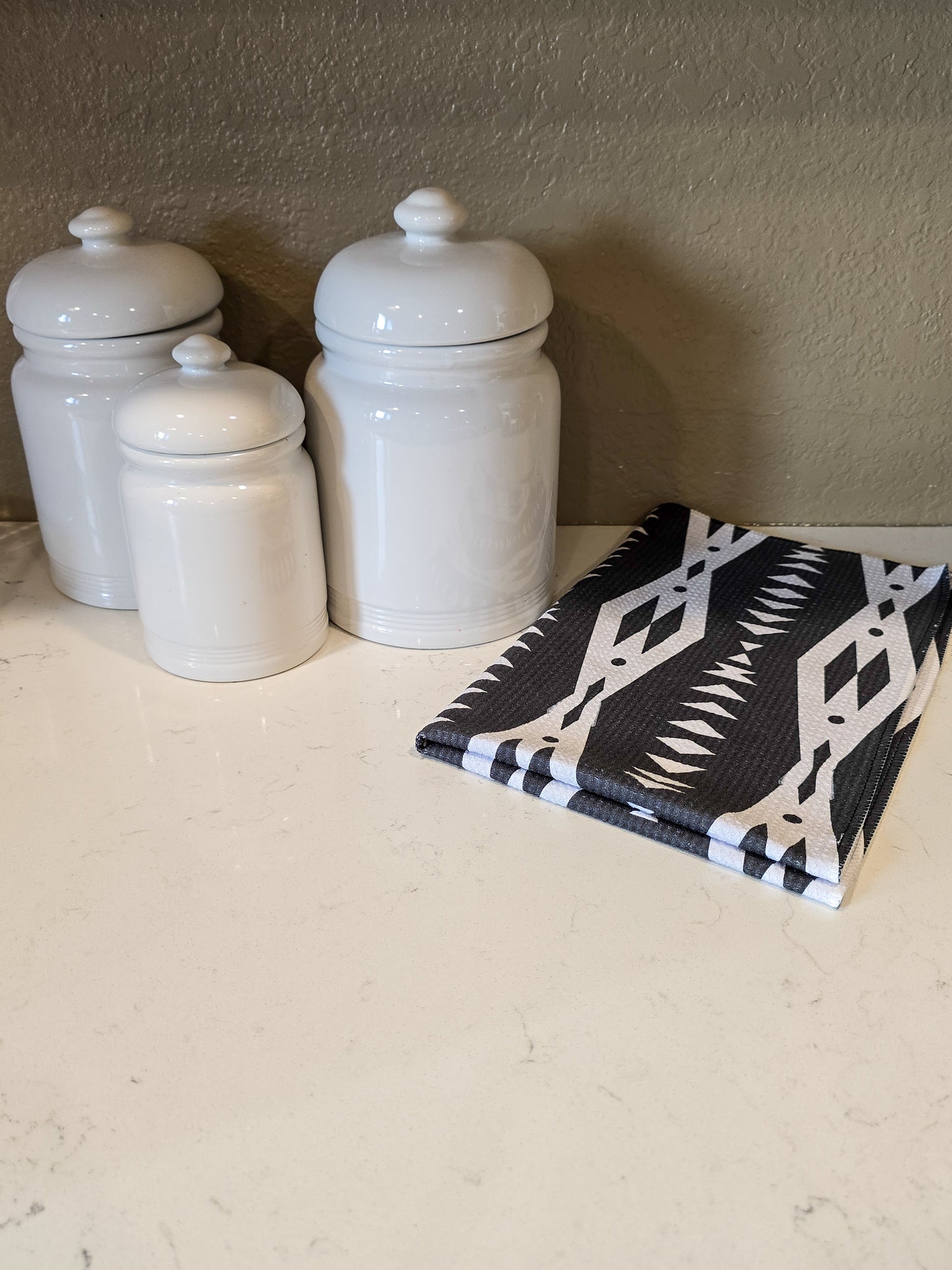Black and white geometric triangle patterned microfiber hand towel folded to just show the front placed on a kitchen counter next to three white canisters. 