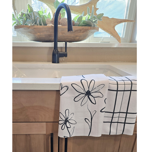 Double-sided waffle weave microfiber hand towel draped over a white kitchen sink, showing a white front with black hand-drawn floral designs and small heart accents, alongside a black grid pattern on the reverse side.