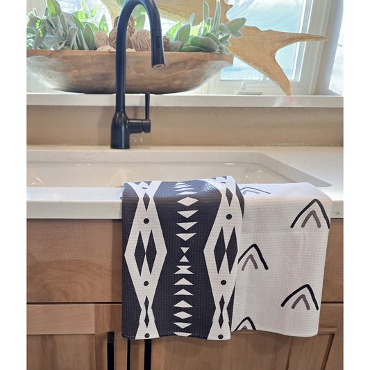 Double-sided waffle weave microfiber hand towel draped over a white kitchen sink, showing a black front with white geometric diamond and triangle patterns and a white reverse side with small black and gray arrow-style triangle designs.