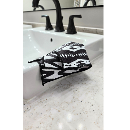 Black and white waffle weave microfiber washcloth folded over the edge of a bathroom sink, showing both patterned sides. The front having geometric shapes and patters, the back having zigzag lines.