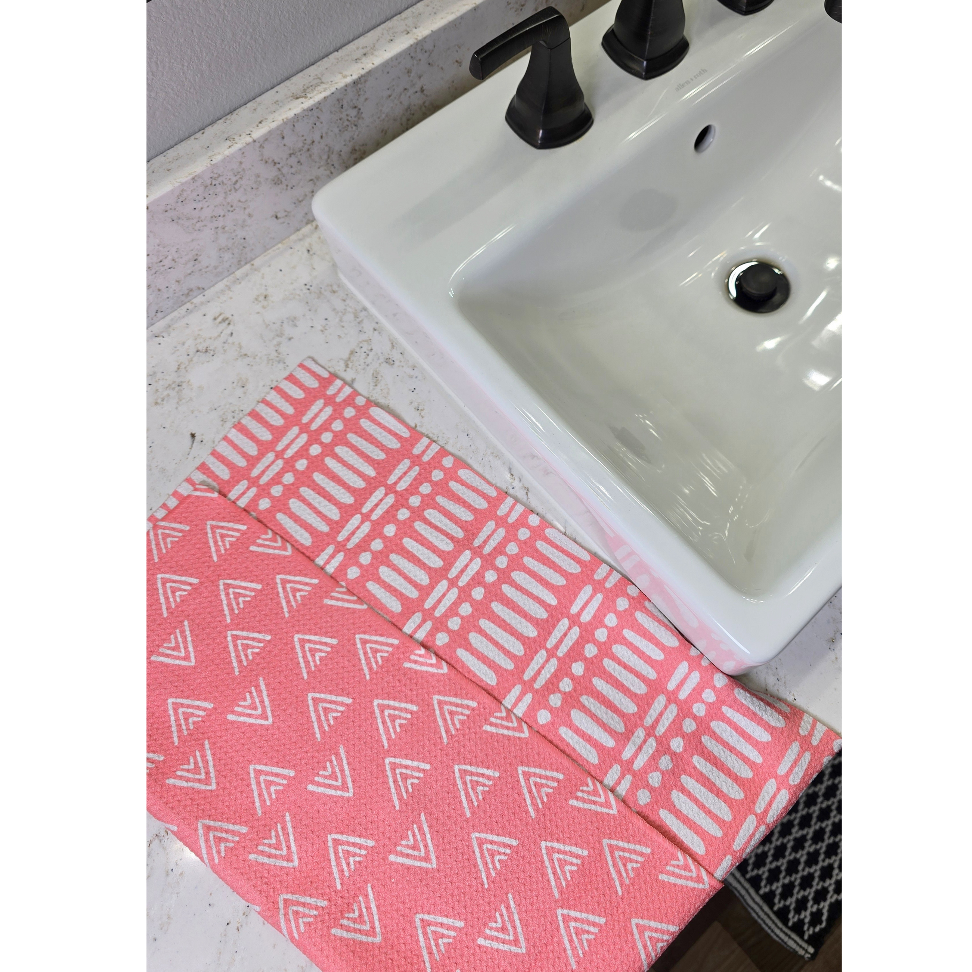Bright pink waffle weave microfiber hand towel with white geometric patterns, folded on the counter next to a white bathroom sink to show both the white triangle patterned front and the coordinating reverse white line and dot patterned side.