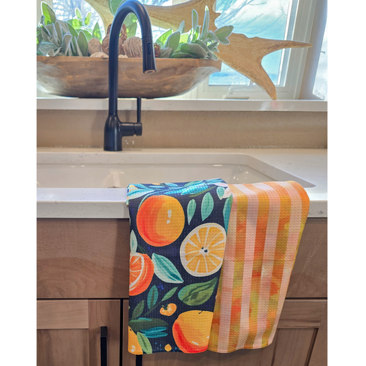 Double-sided waffle weave microfiber hand towel draped over a white kitchen sink, showing a navy front with bright citrus fruit, green leaves, and white floral designs, alongside a reverse side with vertical orange and yellow striped pattern.