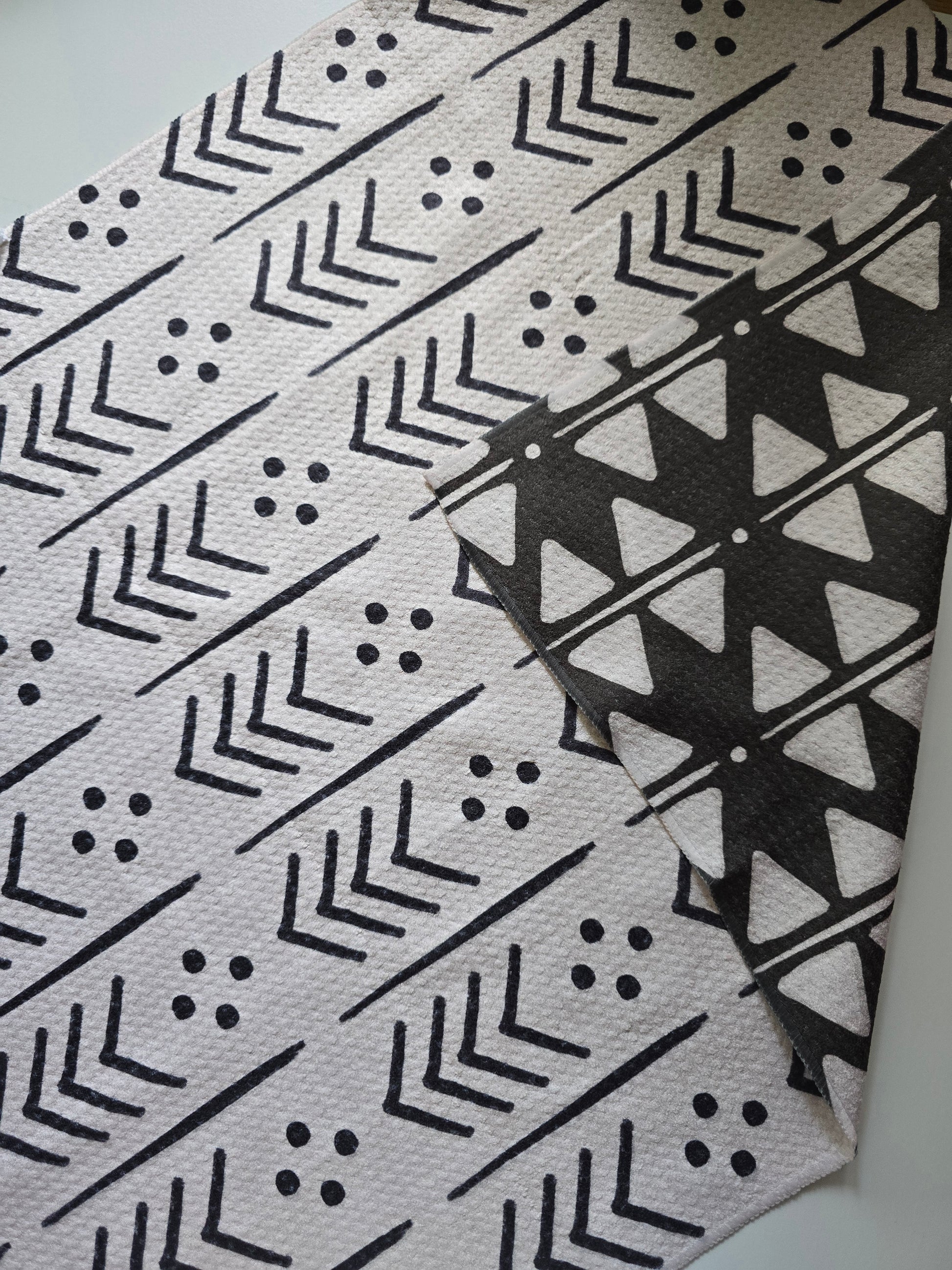 Close-up of a single microfiber towel showing the backside with a corner folded back to show the reverse side. Towel is laying on a white surface. This backside features a cream background with black arrow, line and dot designs, and the corner showing the reverse side shows a black background with white geometric triangle, dot and line patterns.