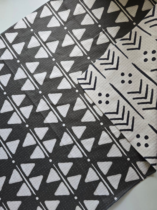 Close-up of a single microfiber towel with a corner folded back to show the reverse side. Towel is laying on a white surface. The front features a black background with white geometric triangle, dot and line patterns, and the back shows a cream background with black arrow, line and dot designs.