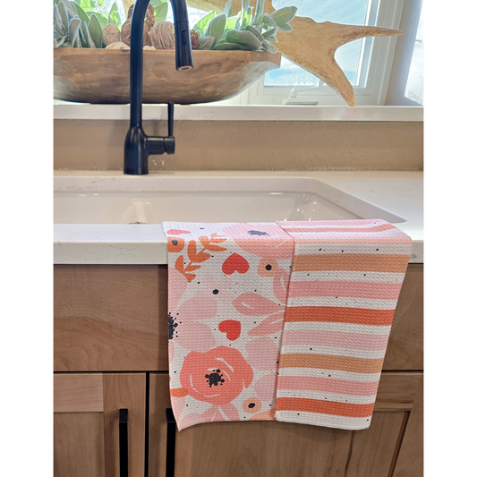 Double-sided waffle weave microfiber hand towel draped over a kitchen sink, showing a floral pattern in soft pinks and corals with black splatter on one side and horizontal peach, coral, soft pink and white stripes with black splatter on the reverse.