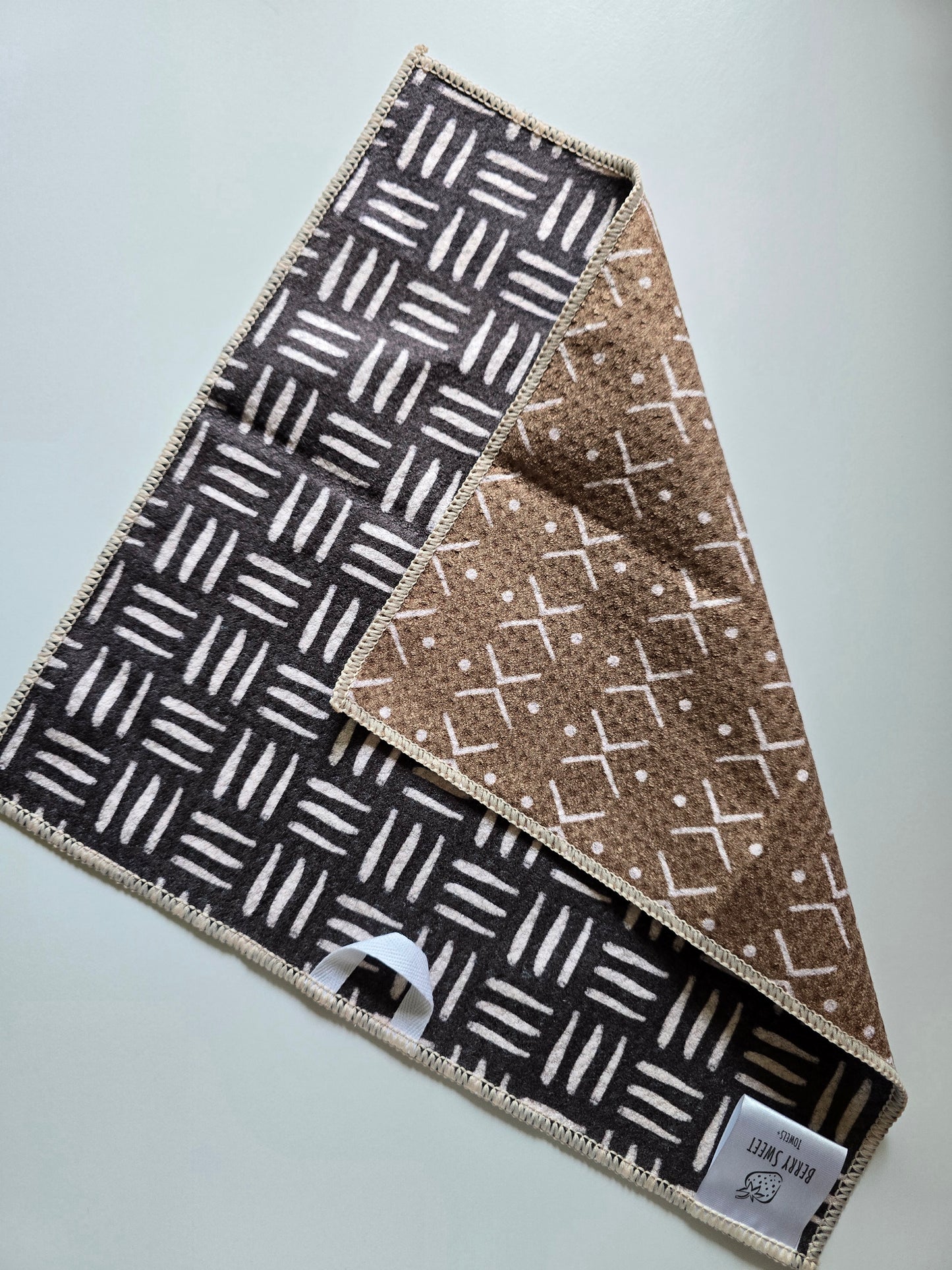 Double-sided waffle weave microfiber washcloth with dark brown side featuring white geometric line pattern and light brown reverse side with white cross and dot pattern, shown partially folded.