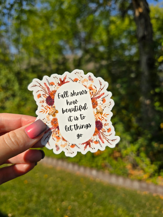 Scalloped-edge vinyl sticker featuring a fall floral wreath in warm orange and brown tones surrounding the quote, “Fall shows how beautiful it is to let things go,” on a white background shown in an outdoor setting with green grass and green trees.