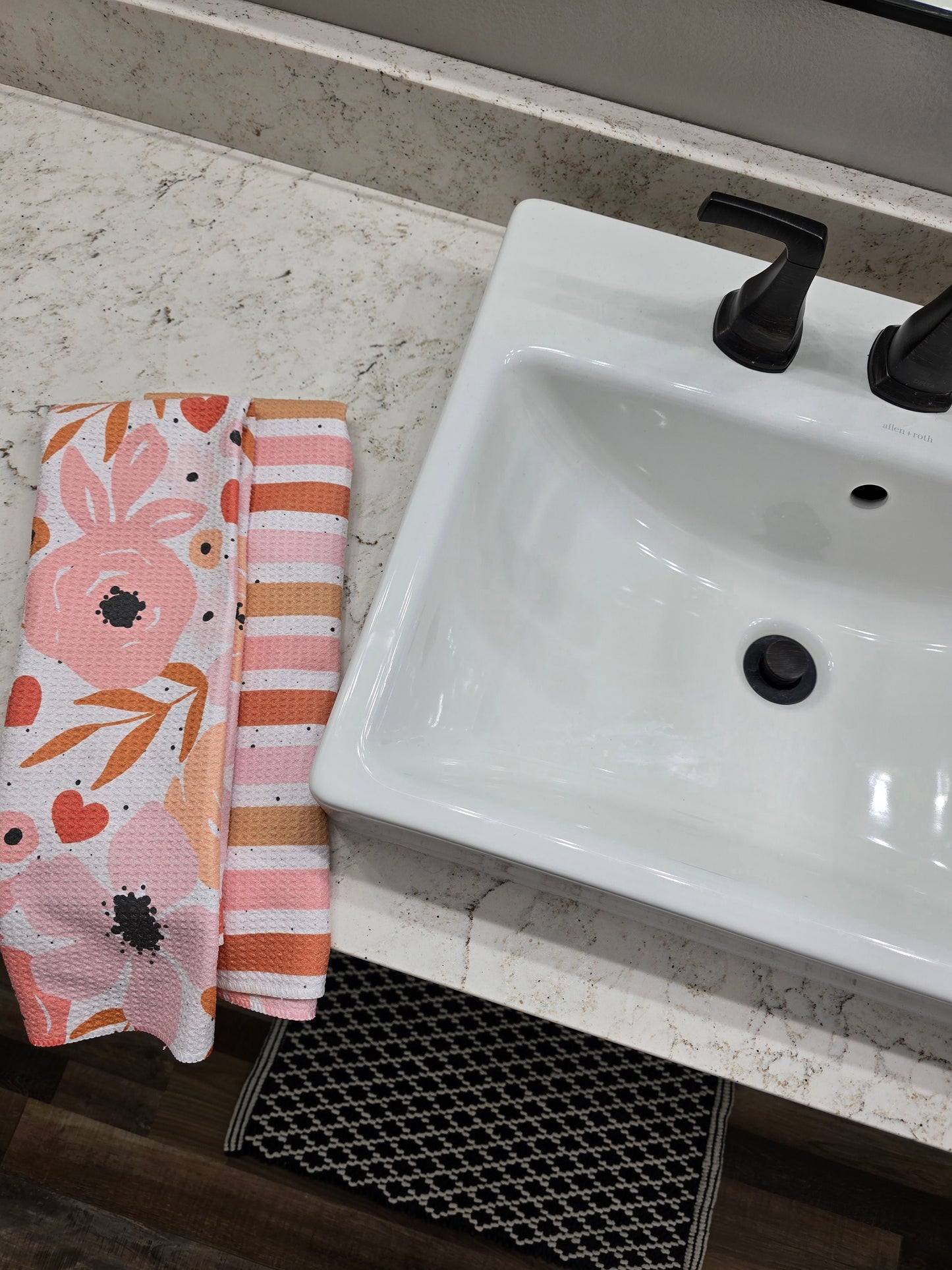Double-sided waffle weave microfiber hand towel folded next to a white sink, showing a floral pattern in soft pinks and corals with black splatter on one side and horizontal peach, coral, soft pink and white stripes with black splatter on the reverse. 