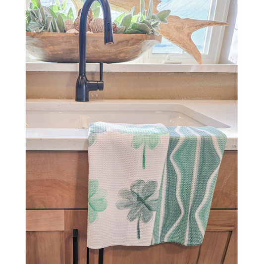 Double-sided waffle weave microfiber hand towel draped over a white bathroom sink, featuring dark and light green four leaf clovers on a white background on the front and a dark and light green and white wavy stripe pattern on the reverse side.