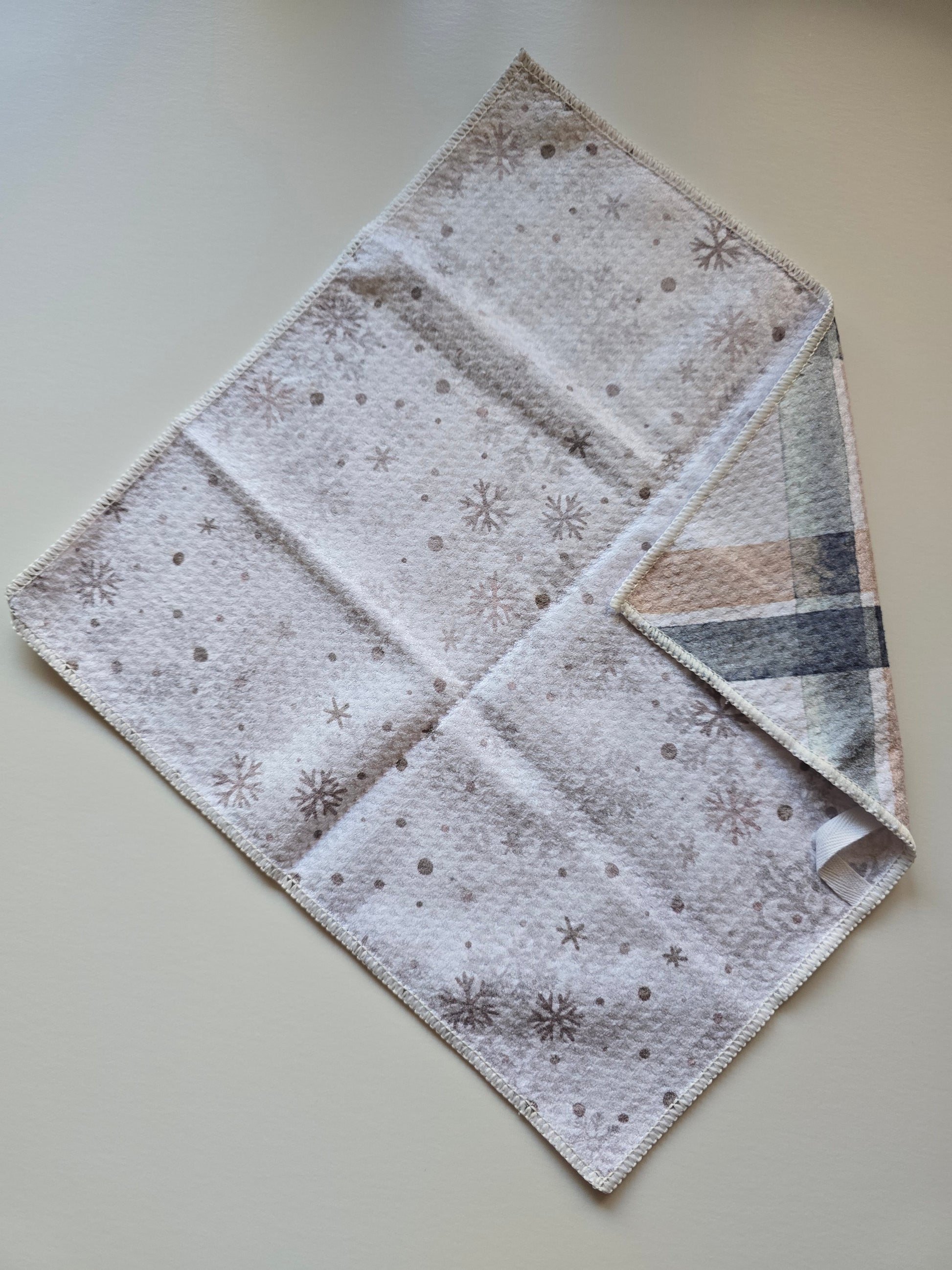 Full view of a waffle weave microfiber washcloth with cream and silver background featuring gold snowflake pattern, with a folded corner revealing the blue and tan plaid reverse side on plain white background. 