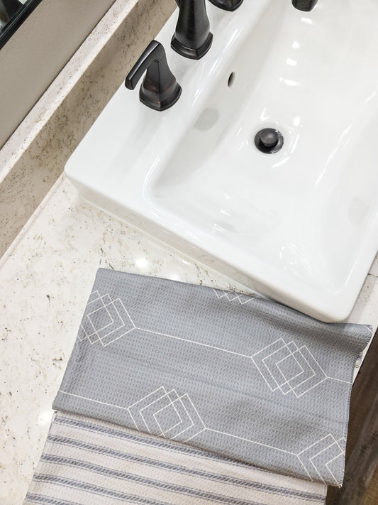 Single waffle weave microfiber hand towel folded to show both sides, with a gray background and white geometric diamond patterned front and a light and dark gray striped reverse, displayed on a bathroom countertop beside a sink.