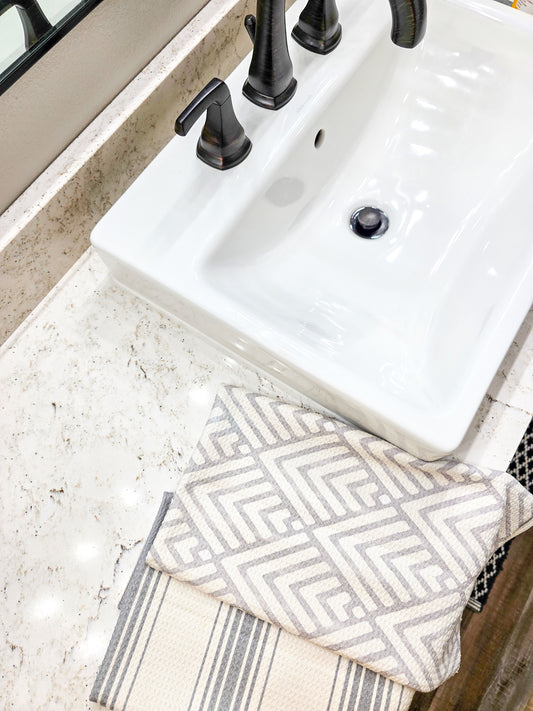 Single waffle weave microfiber hand towel folded to show both sides, with a light gray geometric patterned front on a white background and a gray and white striped reverse, displayed on a bathroom countertop beside a sink.