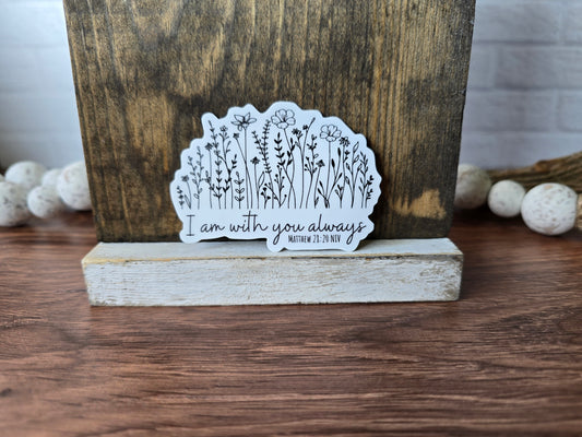 White vinyl sticker featuring delicate black line drawings of wildflowers above the handwritten phrase “I am with you always” with smaller text reading “Matthew 28:20 NIV,” displayed in front of a rustic wood background.