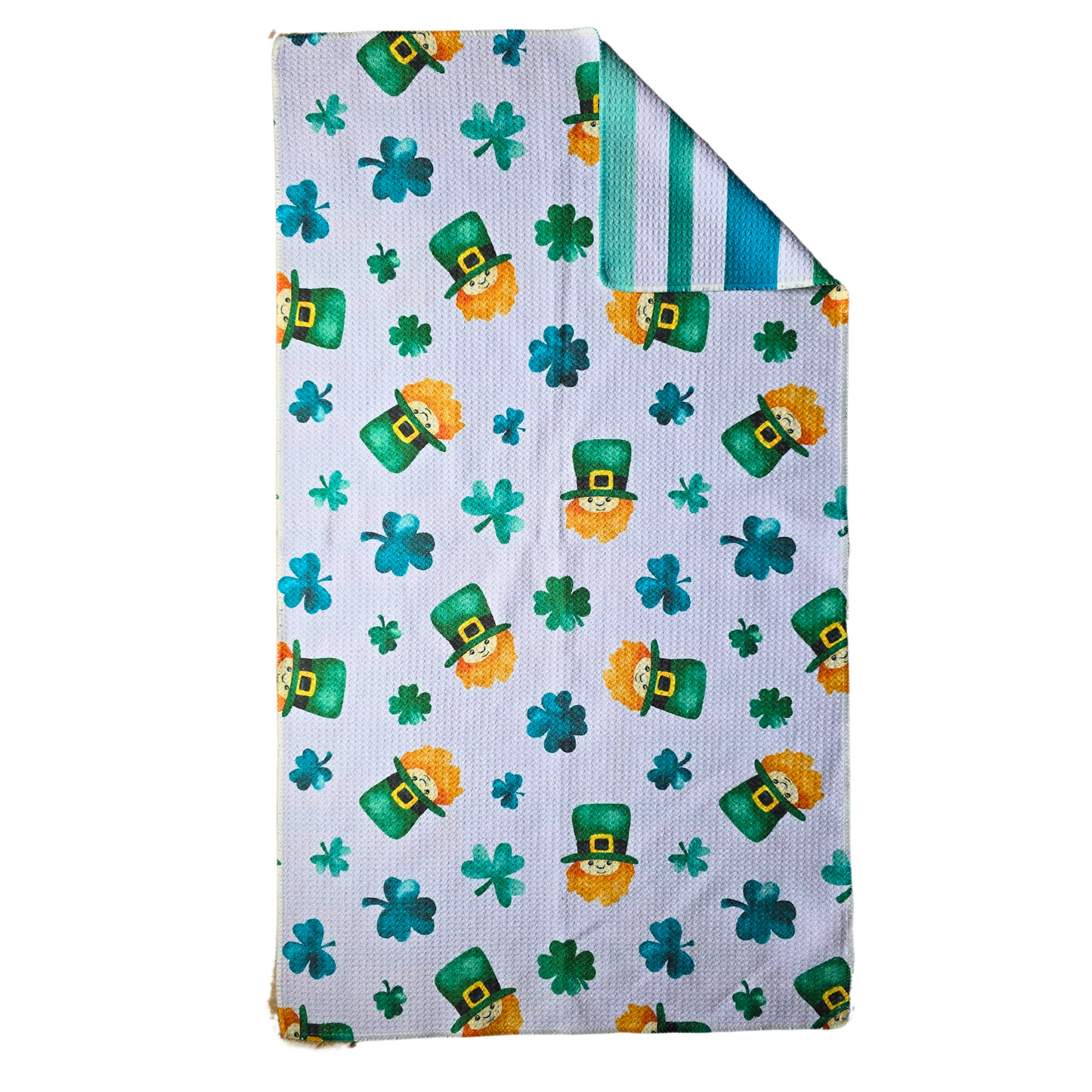 Double-sided waffle weave microfiber hand towel laid flat, showing a white background with repeating various green shamrocks and leprechaun faces wearing green hats with gold buckles, with a folded corner revealing the green and teal striped reverse side.