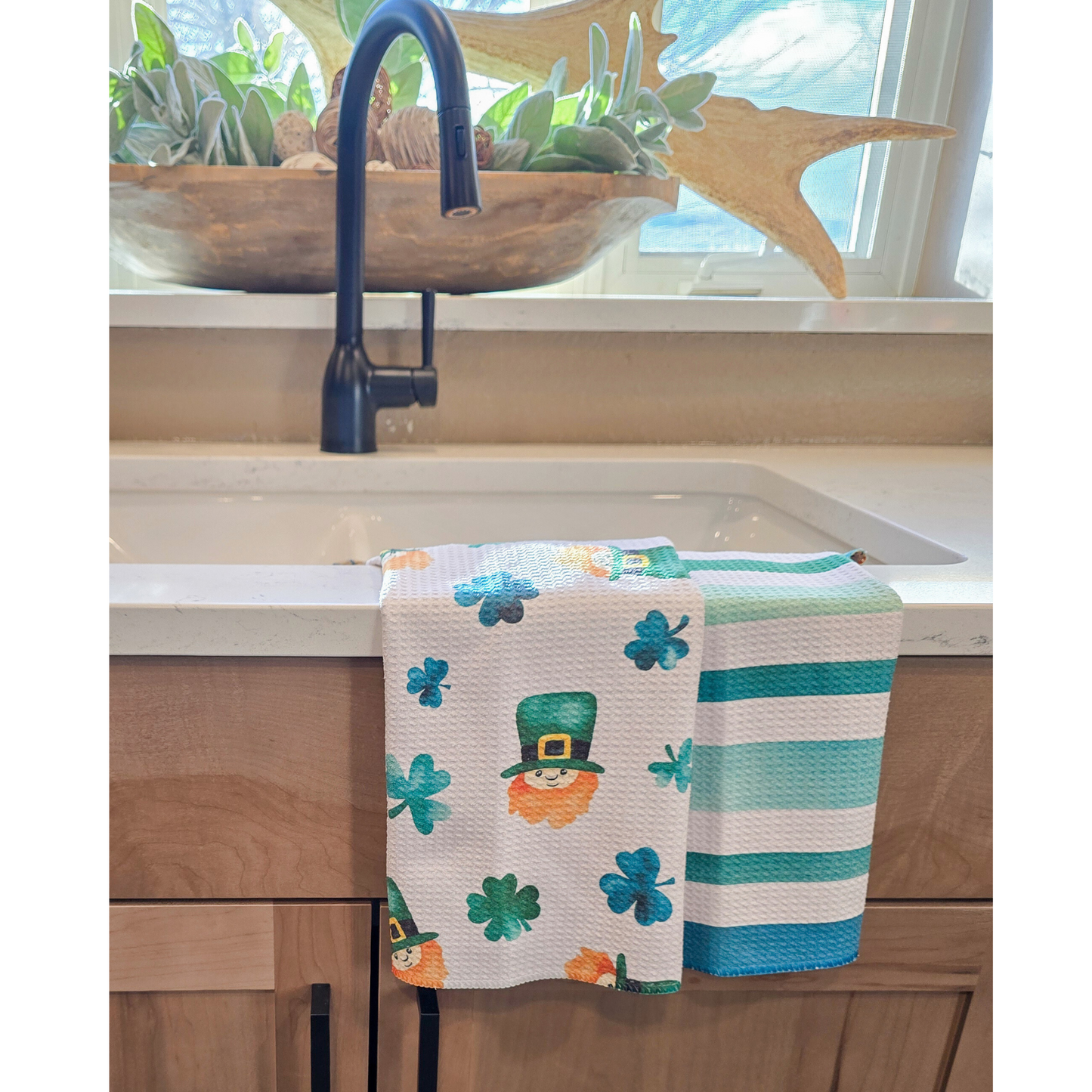Double-sided waffle weave microfiber hand towel draped over a white kitchen sink, featuring various green shamrocks and leprechaun faces with green hats and orange beards on a white background, with green and teal horizontal stripes on the reverse side.
