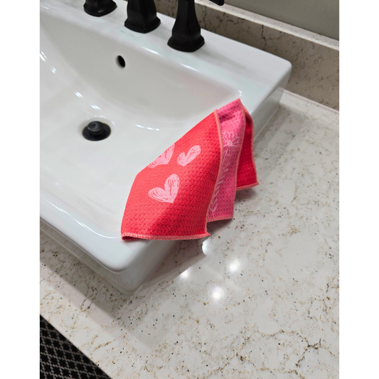 Double-sided waffle weave microfiber washcloth draped over a bathroom sink, showing an orange-pink front with light pink heart pattern and a folded edge revealing the pink reverse side with floral details.