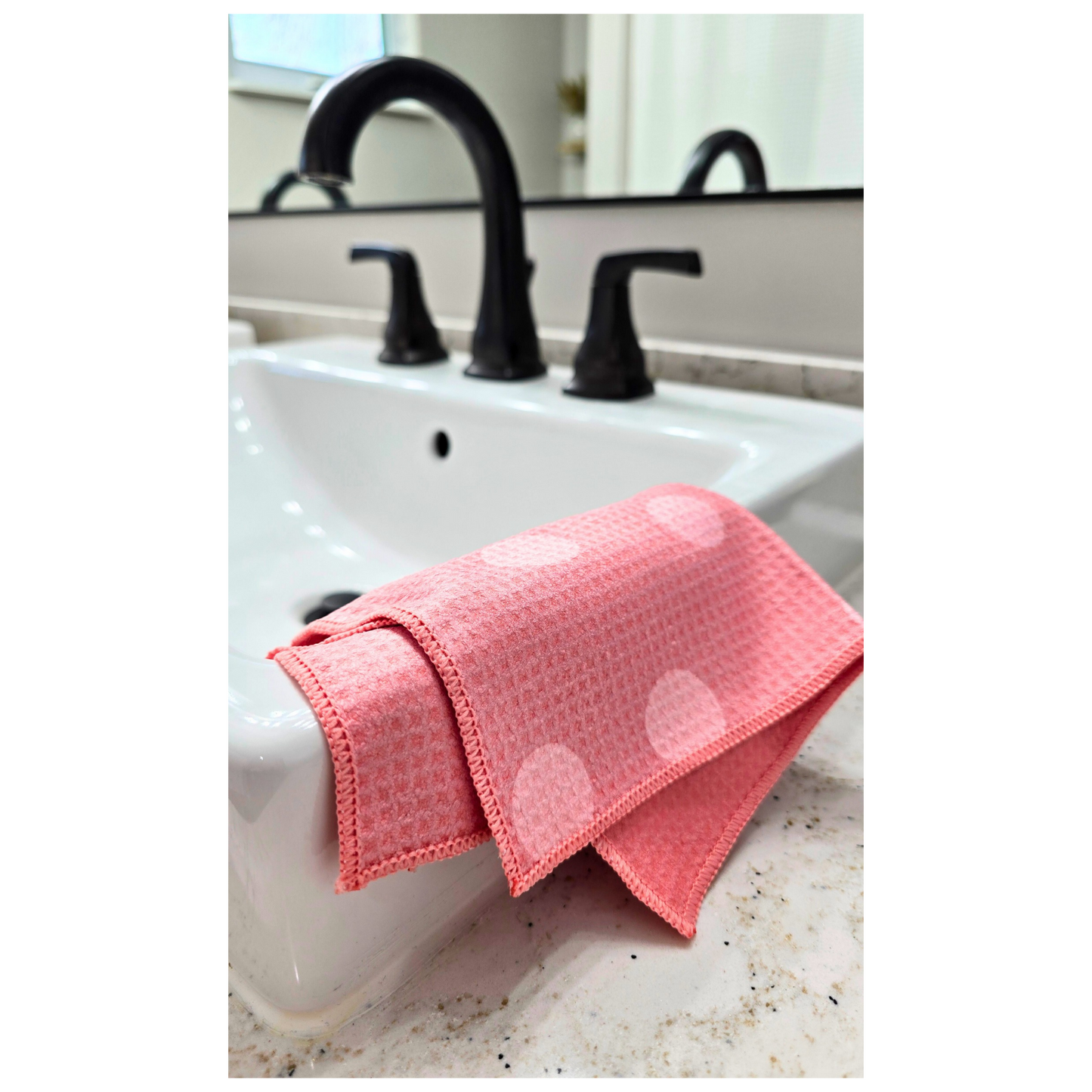Pink waffle-weave microfiber washcloth with light pink polka dots, folded over the edge of a white bathroom sink, also showing the solid pink reverse side.