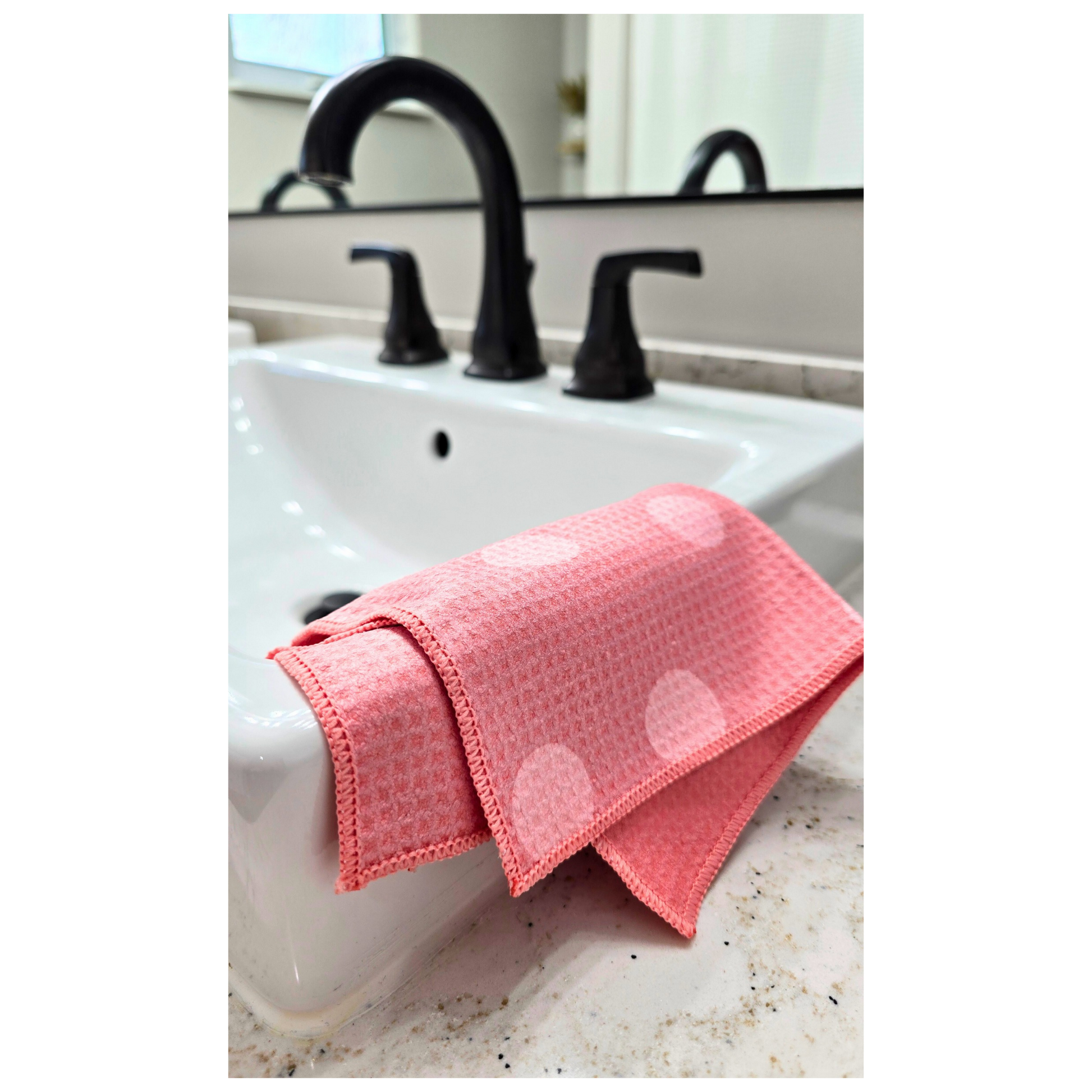 Pink waffle-weave microfiber washcloth with light pink polka dots, folded over the edge of a white bathroom sink, also showing the solid pink reverse side.