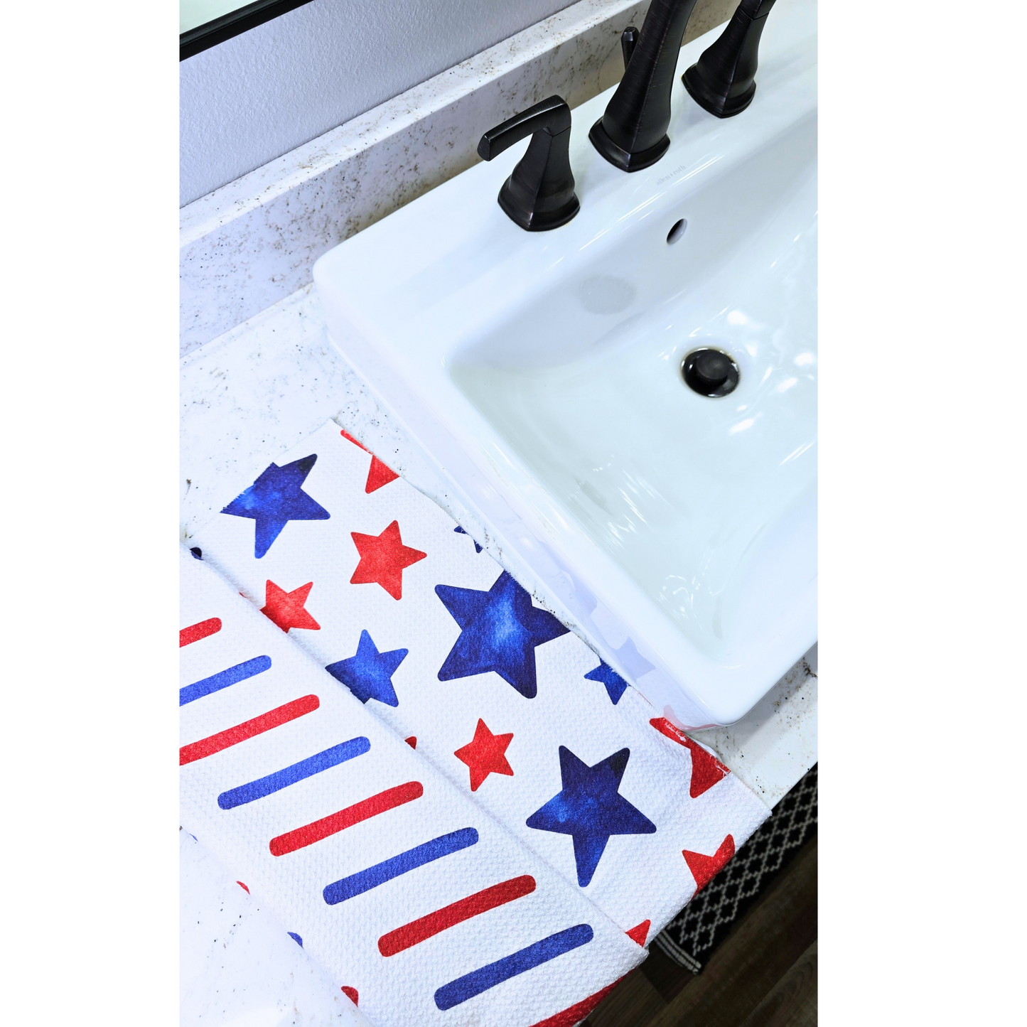 Double-sided waffle weave microfiber hand towel sitting on a white counter next to a white bathroom sink, showing a white front with red and blue star designs and a reverse side with red and blue diagonal stripe pattern.