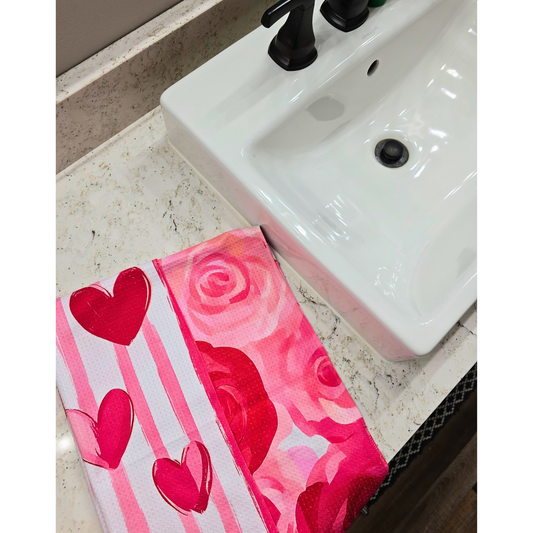 Waffle weave microfiber hand towel folded next to a white bathroom sink, showing a pink and red rose floral front and a reverse side featuring vertical pink and red stripes with heart details.