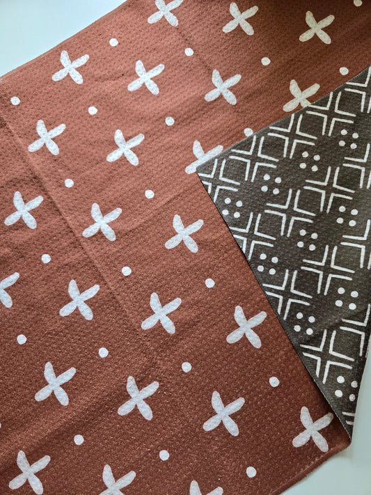 Double-sided waffle weave microfiber hand towel showing rust-colored side with white cross and dot pattern and darker brown reverse side with white geometric pattern.