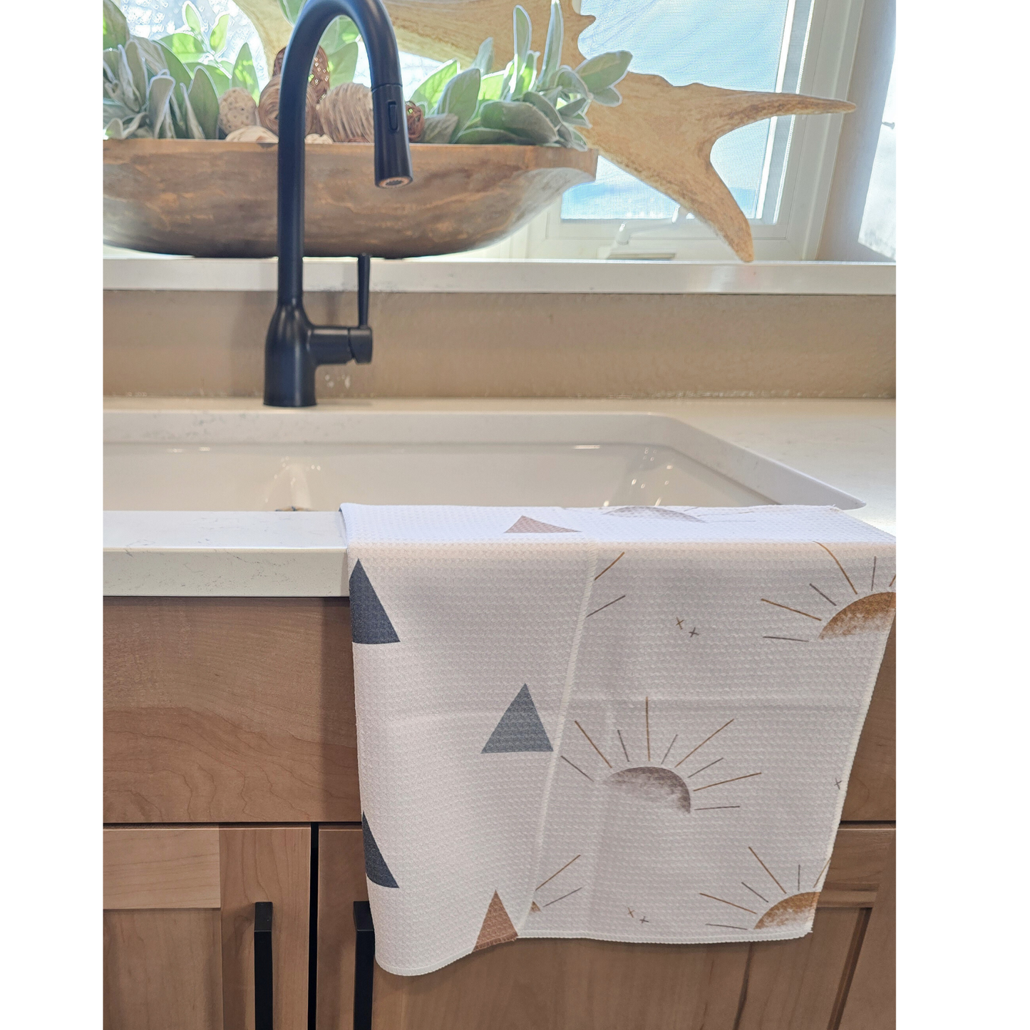 Double-sided microfiber hand towel draped over a white kitchen sink, showing a white front with minimalist sunburst designs in gold and gray, and a reverse whitw side with small geometric triangle patterns in muted blue, gray, and gold.