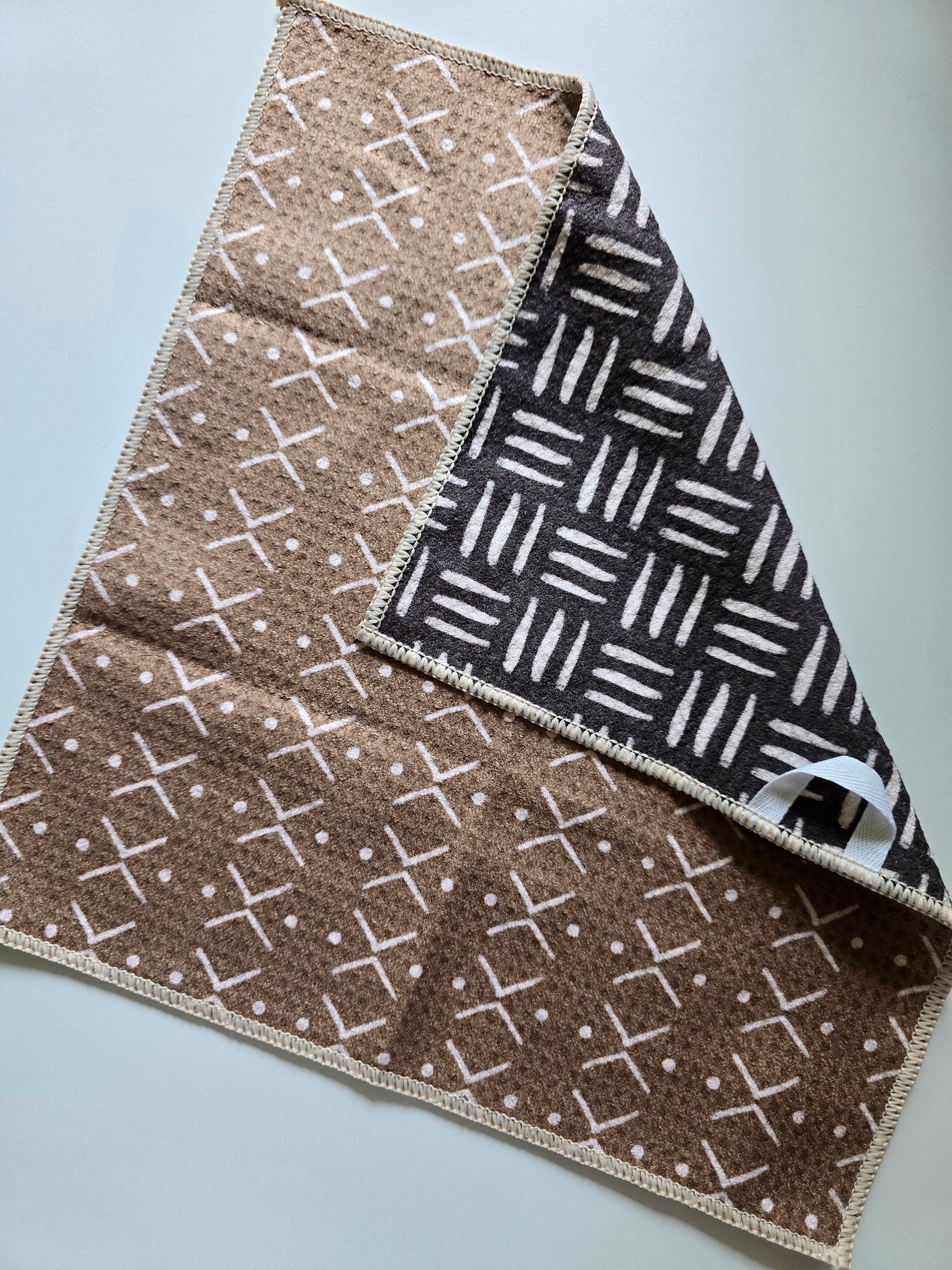 Double-sided waffle weave microfiber washcloth with light brown side featuring white cross and dot pattern and dark brown reverse side with white geometric line pattern, shown partially folded.