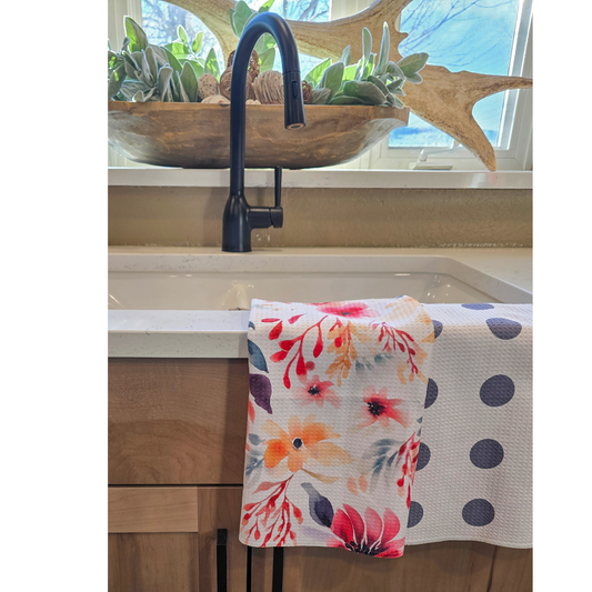 Double-sided waffle weave microfiber hand towel draped over a white kitchen sink, showing a white front with colorful watercolor-style floral designs and a reverse side with evenly spaced purple polka dots on a white background.